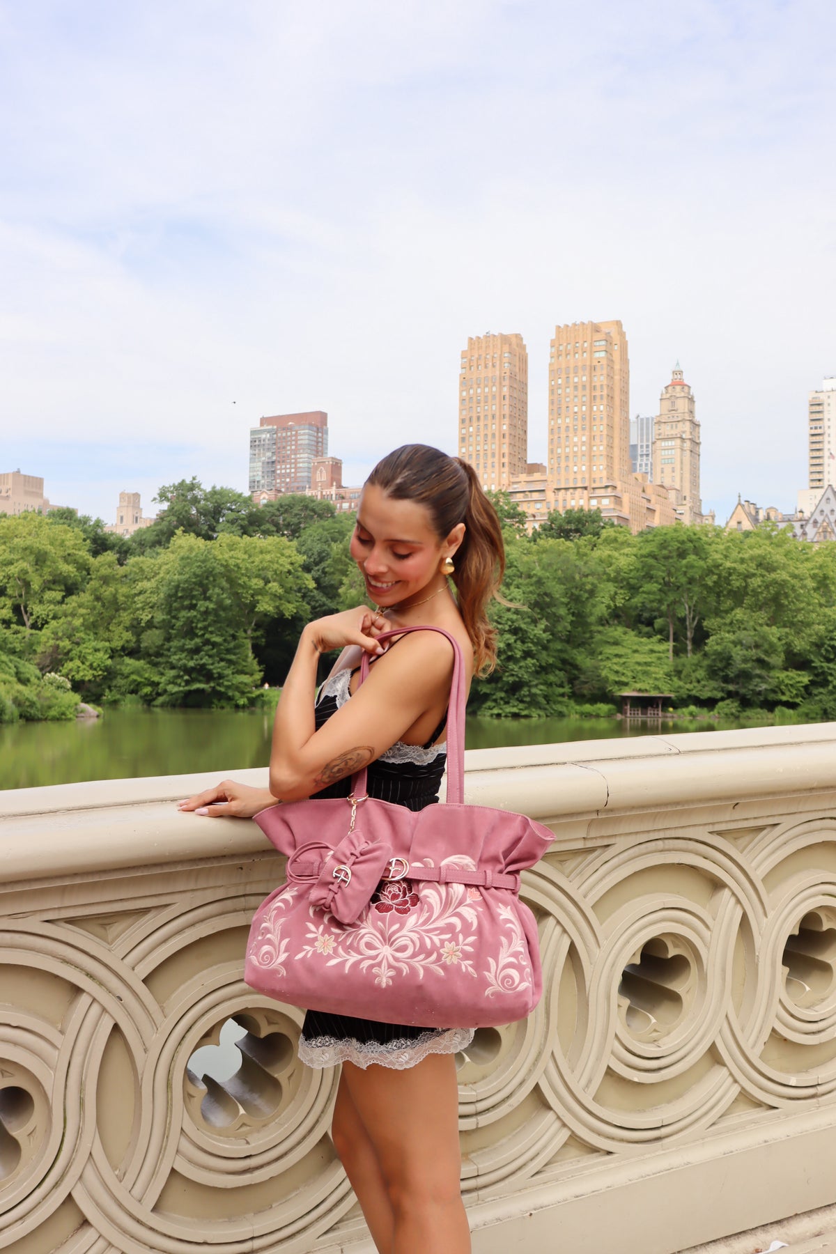A woman posing on a stone bridge in Central Park, New York City, carrying a pink floral embroidered tote bag.