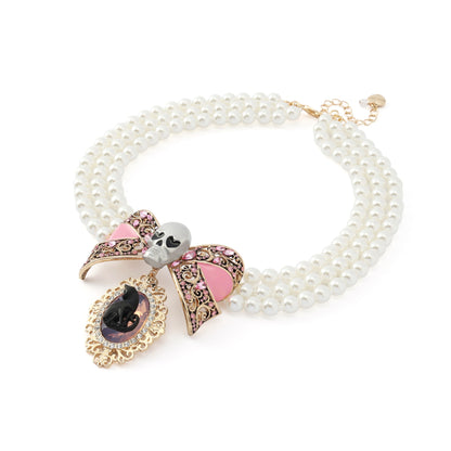 A double-strand pearl necklace with a gothic bow and skull pendant. The necklace features white faux pearls, a gold-tone clasp, and a central pendant decorated with a pink and black bow, a white skull, and a teardrop-shaped black gemstone.
