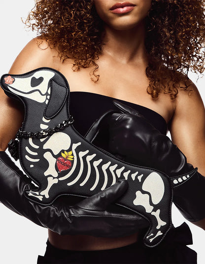 A model in a black strapless top and matching black gloves holds a black faux-leather clutch shaped like a dachshund dog skeleton with a small red heart and yellow flower accent.