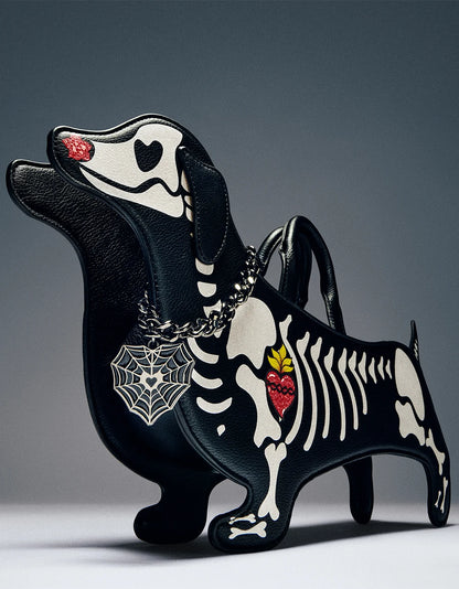 A black leather dachshund-shaped handbag with a skeleton motif, featuring white bone details, a red heart on the chest, a silver spider web pendant, and a red heart on the nose, set against a dark gray studio background.
