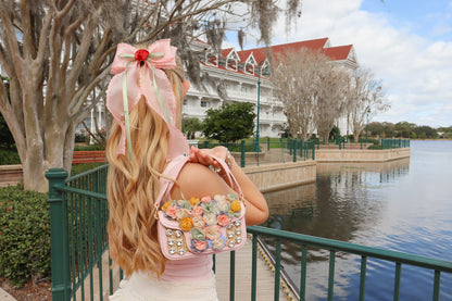 Person with a decorative headpiece and floral bag standing by a waterfront with a building in the background