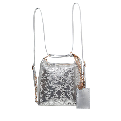 A silver metallic bucket bag with white embroidered lace detailing, featuring a gold chain strap, a matching silver leather hangtag, and a thin shoulder strap, shown against a plain white background.