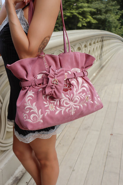 A woman carries a dusty pink fabric shoulder bag with white floral embroidery, a large bow detail, and a gold-tone chain strap, photographed outdoors on a light-colored bridge.