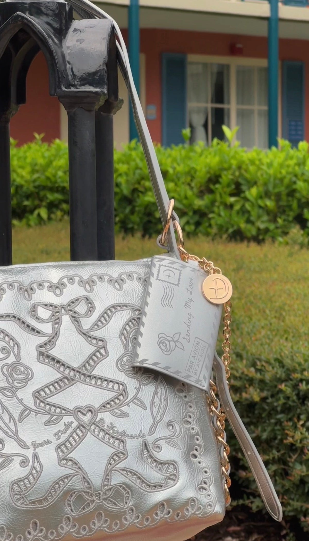 A silver metallic embossed leather shoulder bag with a matching wristlet strap and gold-tone chain, shown outdoors next to a black metal stand with a house and greenery in the background.